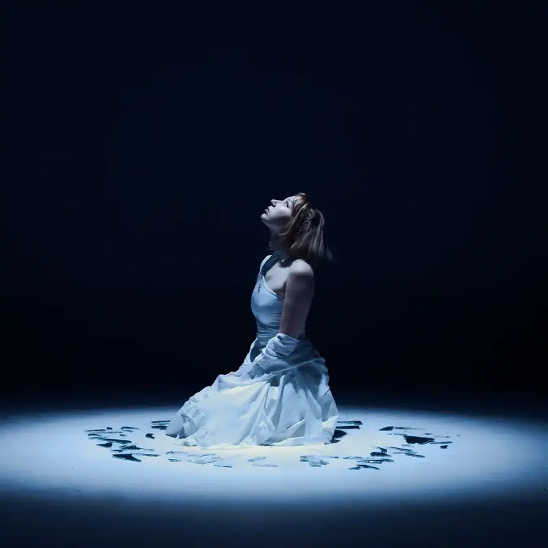 Djouher, dressed in white, kneels in a circle of blue light on a dark stage and looks up.